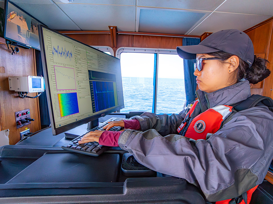 A woman wearing a life jacket in a boat cabin works with complex scientific data on a computer, the ocean stretches to the horizon outside the window