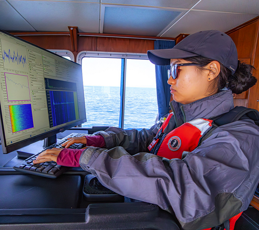 An asian woman wearing a life jacket in a boat cabin works with complex scientific data on a computer, the ocean stretches to the horizon outside the window
